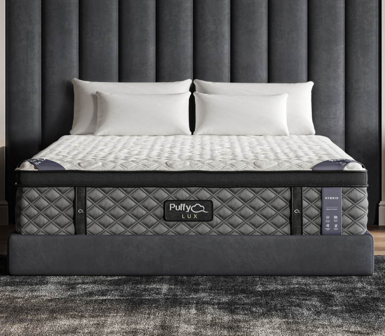 A Puffy Lux Hybrid Mattress with a quilted white cover and grey sides, viewed head-on against a gray upholstered headboard.